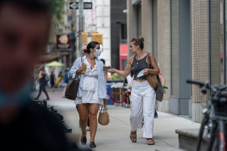 two women talking and walking on the sidewalk, one with a mask on