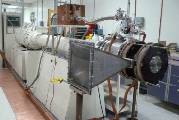 a machine in a laboratory with a rectilinear funnel-shaped structure in the foreground and a long metal pipe receding into the background
