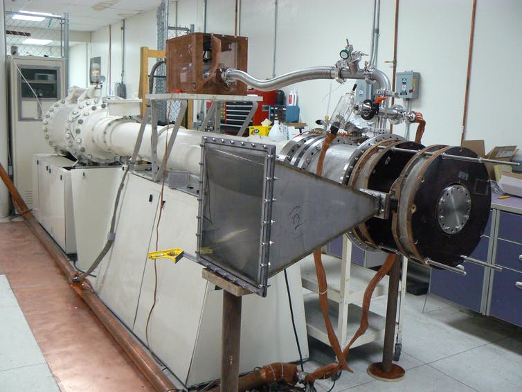 a machine in a laboratory with a rectilinear funnel-shaped structure in the foreground and a long metal pipe receding into the background