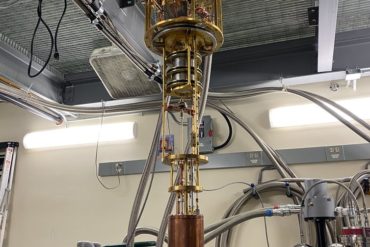 The HAYSTAC detector, a large copper cylinder connected to a gold-plated assembly of tubes and wires hanging from the ceiling of a lab.