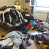 A room with clothes on the floor and piled high on the bed.