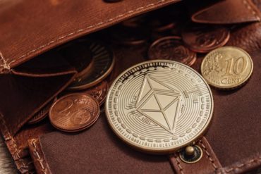 A commemorative coin bearing a double-pyramid logo lies in an open leather wallet containing euro coins