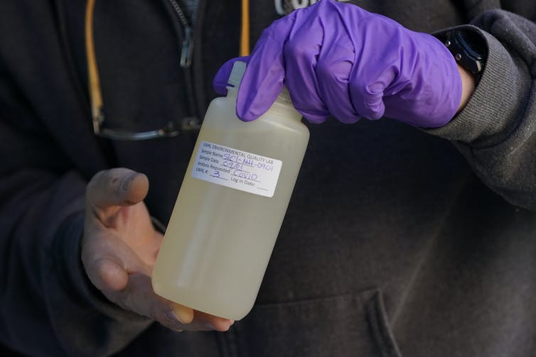 A gloved hand holds a sealed plastic bottle of murky-looking water.