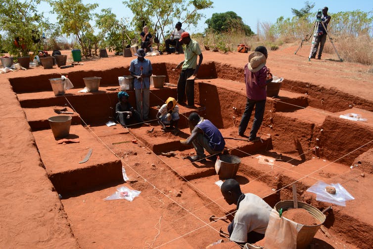 people digging in red earth at an outdoor archaeological site
