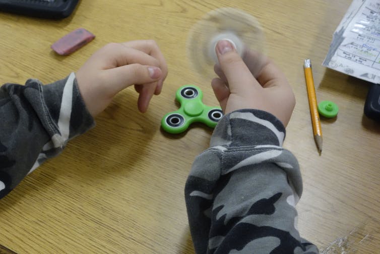 arms of a child wearing camo sweatshirt and holding a white fidget spinner at a school desk with pencil, eraser and green fidget spinner on desk in background