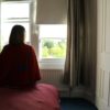 view from behind of woman sitting on bed and looking out window