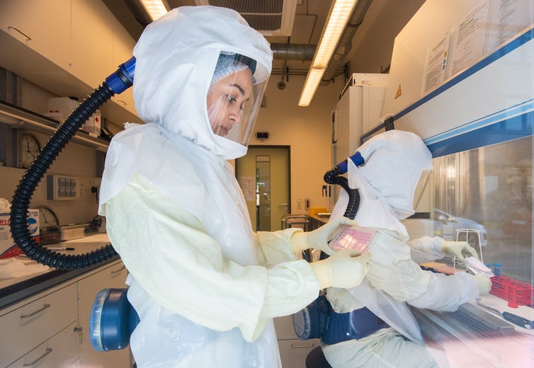 Two scientists in a COVID-19 research laboratory, wearing fully enclosed white protective suits with clear face screens and independent air supplies.