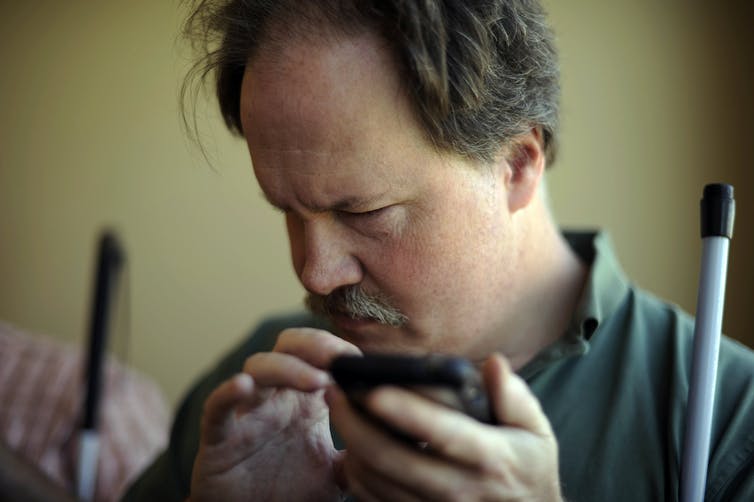 A man holds a smartphone, his face turned away from the screen, a white cane resting against his shoulder
