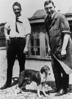 two men in early 20th C clothes standing with a dog between them