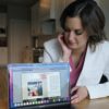 A woman in a white lab coat sitting in a kitchen points a laptop screen to the viewer