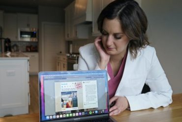 A woman in a white lab coat sitting in a kitchen points a laptop screen to the viewer