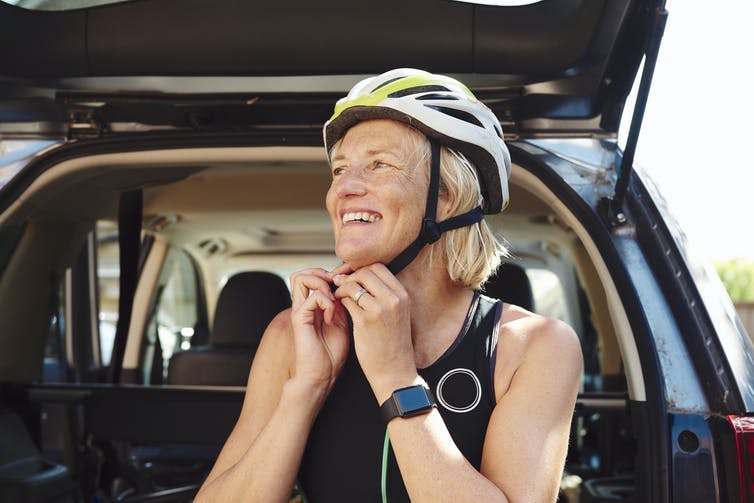 A gray-haired woman snaps on a bike helmet. She is wearing an Apple watch.