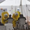 Health care workers in yellow protective equipment treating a group of people in a white tent.