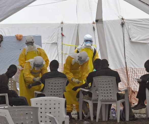 Health care workers in yellow protective equipment treating a group of people in a white tent.
