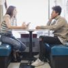 Two teenagers sit across from each other at a table in a cafe. One of them is speaking and the other one is listening.