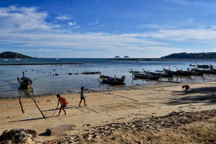 Moken children play on the beach, with small boats tied up in the shallows