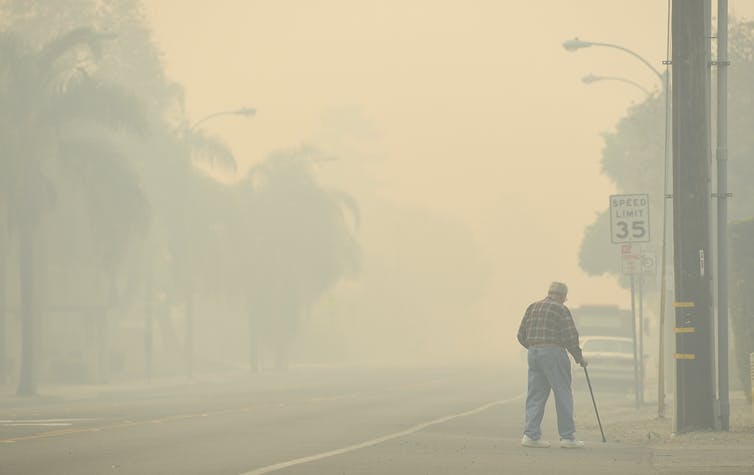 An older man with a cane crosses n empty road in smoky conditions.
