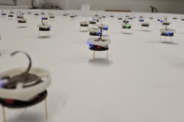 Several dozen small plastic discs containing electronics and perched on metal wire legs  are spread across a smooth featureless surface