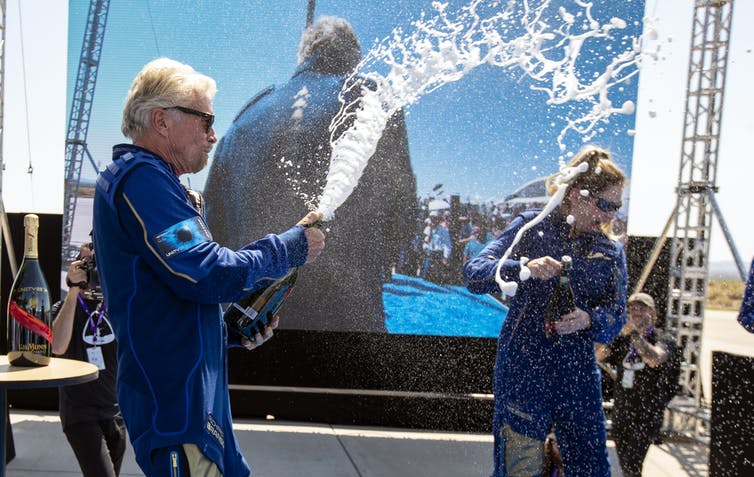 Richard Branson in a blue jumpsuit celebrated his successful sub-orbital flight to space by spraying champagne on a stage.