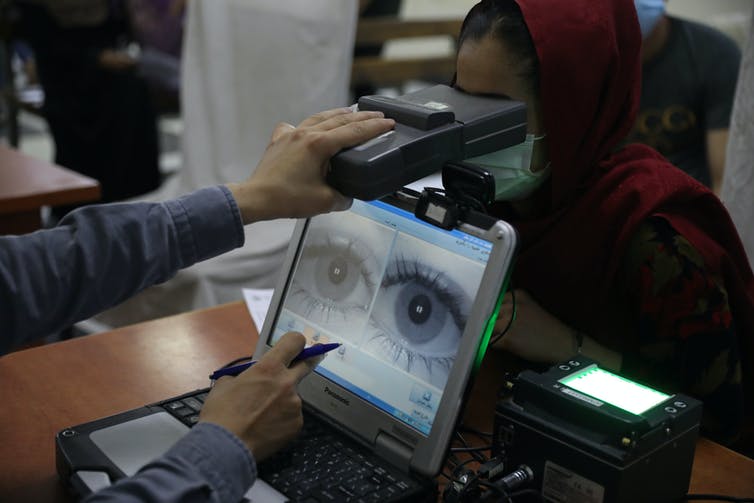 A computer screen shows an enlarged image of a pair of eyes as an arm holds a boxlike object in front of the eyes of a woman wearing a headscarf and facemask