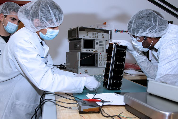Three people in white lab coats and hairnets working on a satellite roughly the size of a loaf of bread.