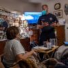 A man in a blue uniform who is wearing a mask speaks to a woman sitting in a recliner in a living room.