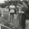 Black and white photo of a woman speaking to six men gathered around a solar panel.