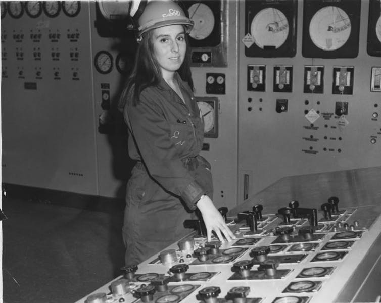 Black and white photo of a woman in a hard hat at the controls in a large control room.