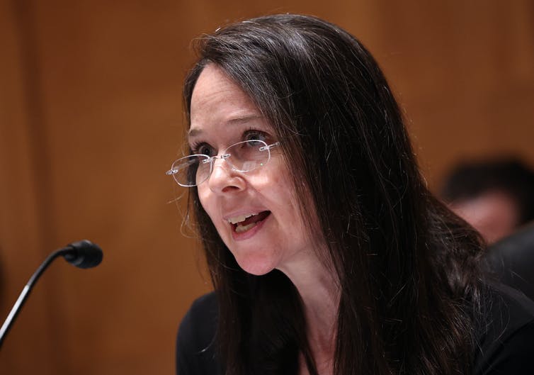 a woman with long dark hair wearing eyeglasses speaks into a microphone