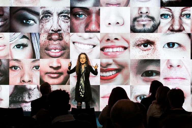 A woman stands on stage in front of an audience gesturing with her hands as the screen behind her displays a mosaic of close-up images of parts of people's faces