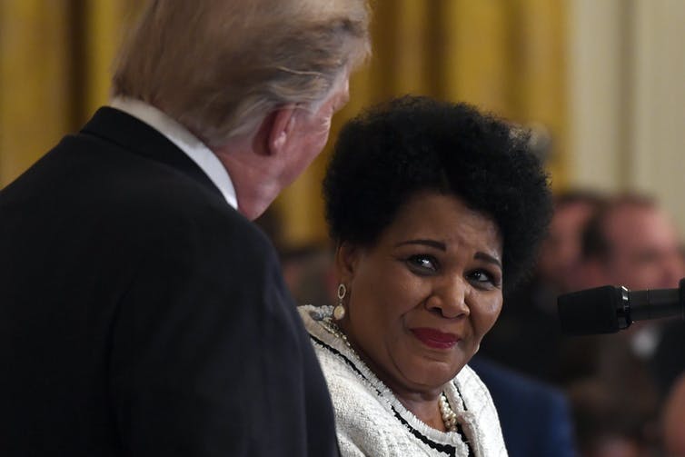 A woman in a white suit looks up at a man in a suit with his back to the camera.