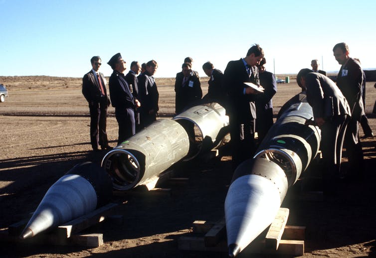 a group of men look at a pair of disassembled missiles lying on the ground in a desert