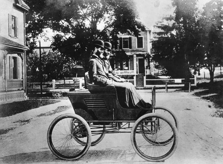 Two grown identical twins with beards, wearing bowler derbies, atop an early automobile