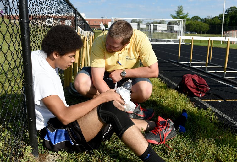 A young player sits against a fence next to a track looking exhausted while a man crouches down next to him and talks to him.
