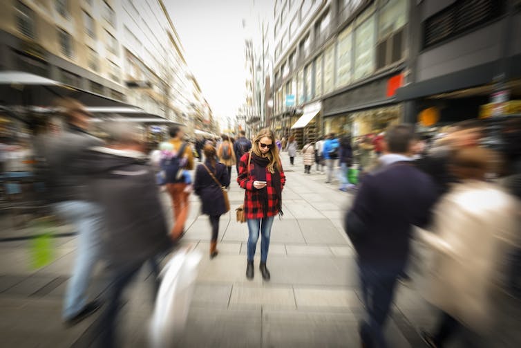 a city street view with a young woman looking down at her phone in focus while passersby are out of focus