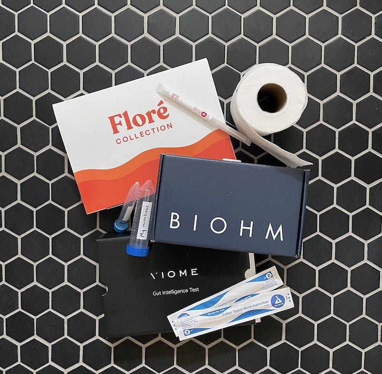 Three gut microbiome test kits (Floré, Biohm, Viome) and a roll of toilet paper displayed on a tile floor