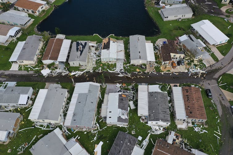 Rows of closely spaced homes next to an inlet or lake. Several have clearly damage roofs.