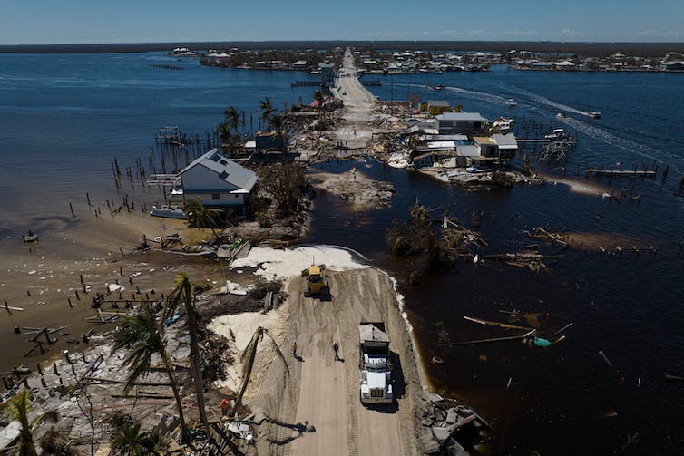 A damaged narrow roadway with water on both sides and damaged homes.