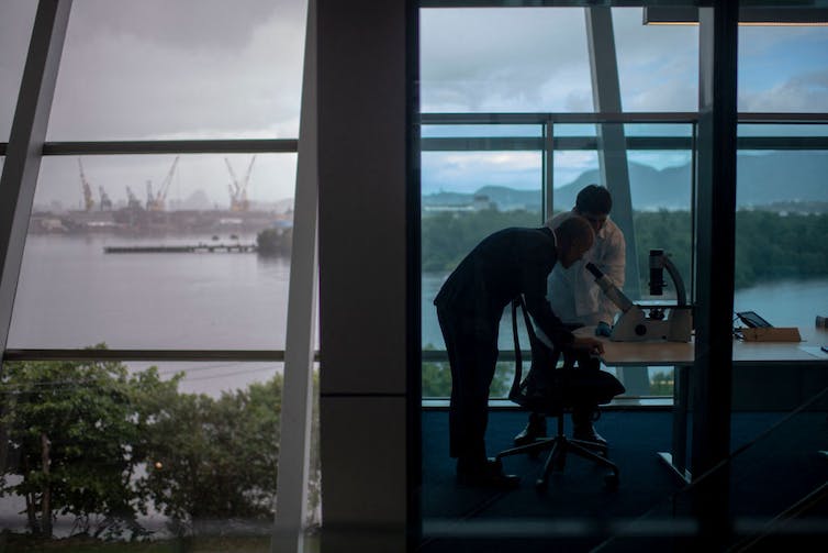 Two men bend over a microscope in an office with big glass walls overlooking water.