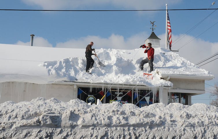 Two people shovel knee-deep snow off a roof.