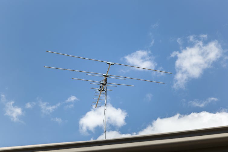 Against a blue sky and white clouds, a TV antenna sits on top of a house.