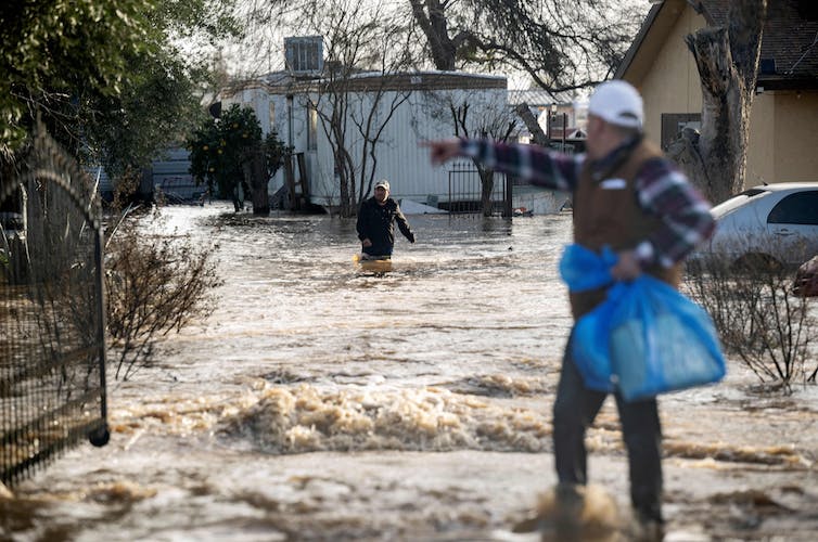 A man carrying a plastic bag gestures to someone in knee-deep floodwater near a home.