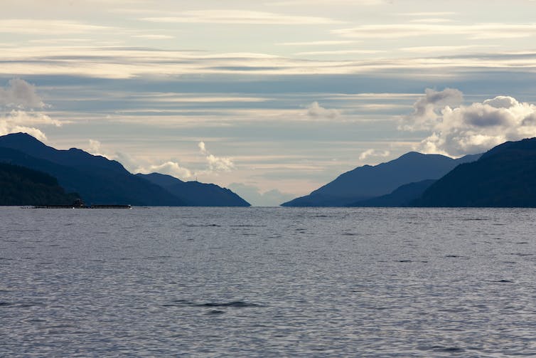 The photograph shows a blue sky, white clouds, highlands and the murky waters of Loch Ness.