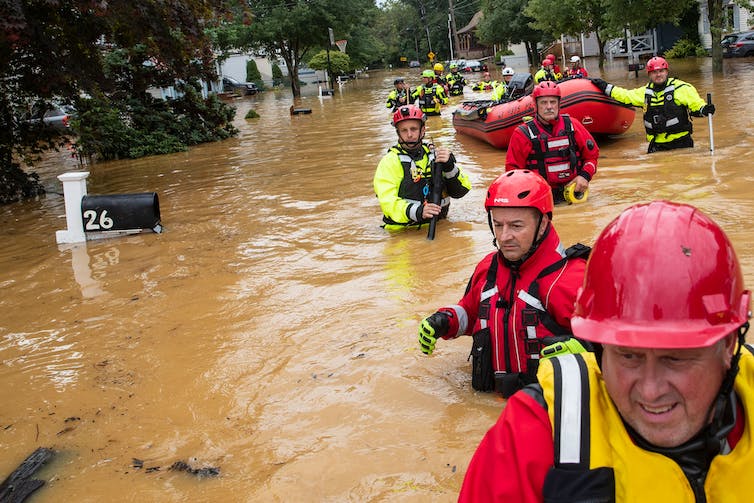 A line of rescue workers in bright vests and hard hats walks in waist-deep water on a flooded street, pulling a raft. Water is up to the mailbox they're passing.