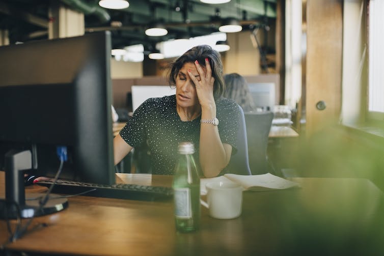 a woman seated at a desk in front of a computer with her eyes closed and her left hand on her forehead