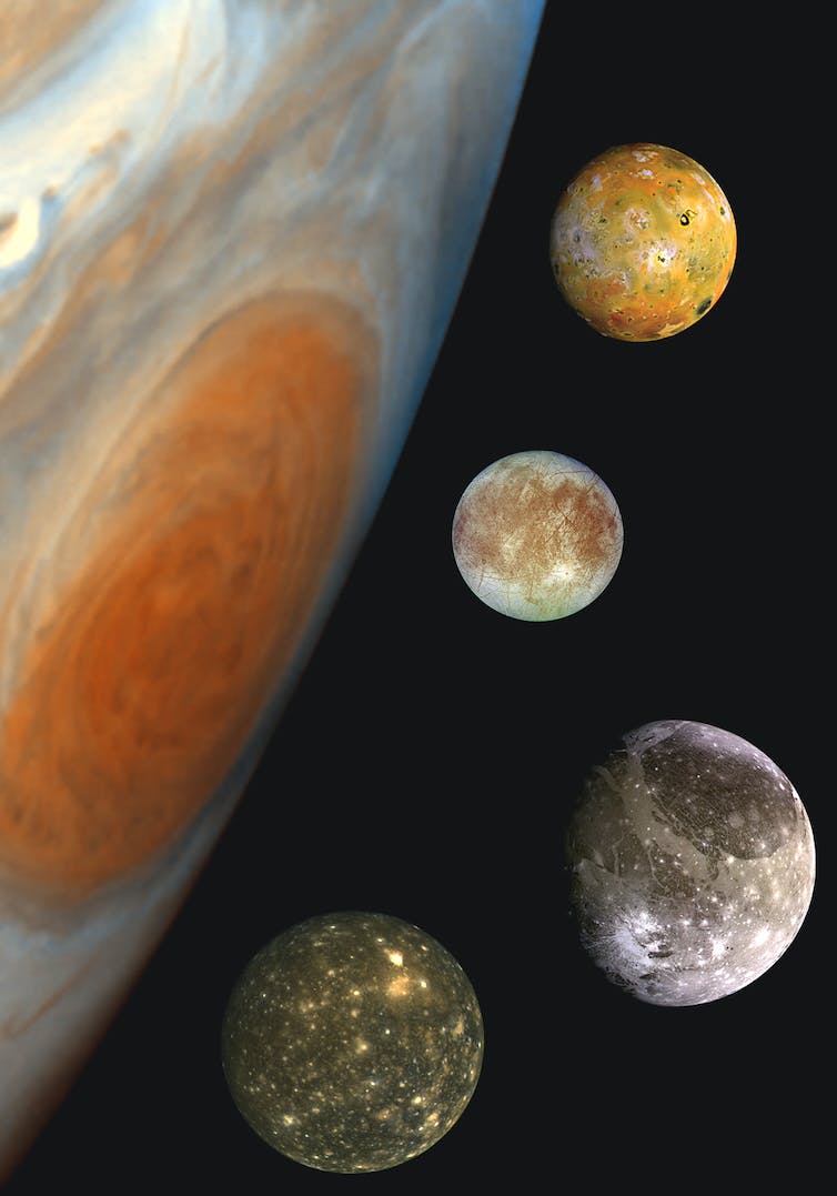 Four moons next to a large red spot on the surface of Jupiter.