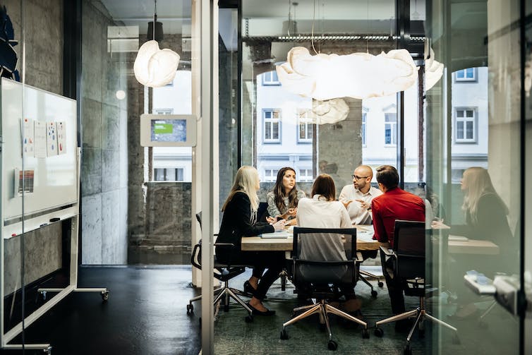 Six people sit at a table in a brightly lit office with pie charts on the wall and windows in the background.