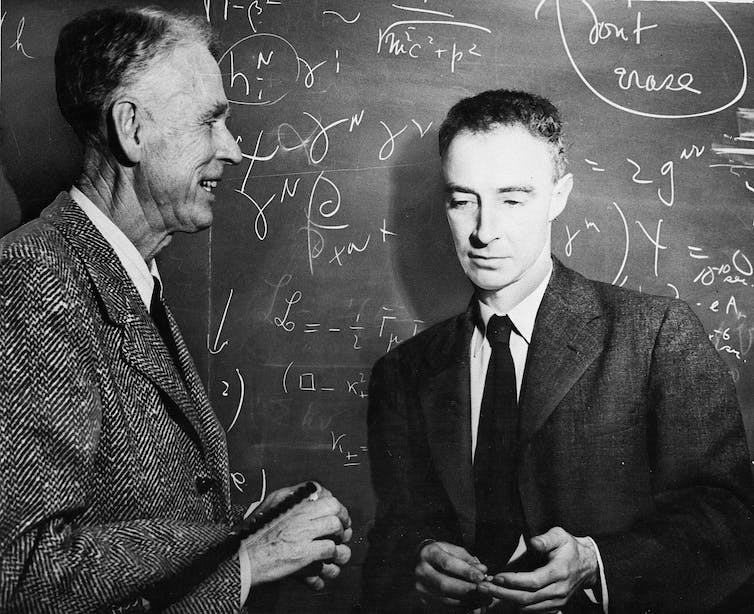 A black-and-white old photo of two men wearing jackets and ties. The one on the right is younger and looking down, in the backround is a blackboard with equations written on it.