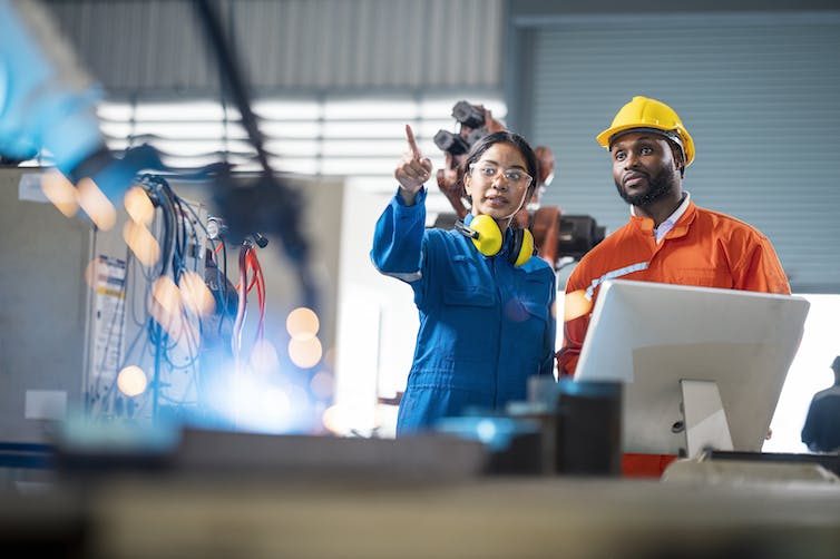 Two engineers standing in a factory, the one on the left, a woman, is pointing, while the one on the right, a man, wears a hard hat.