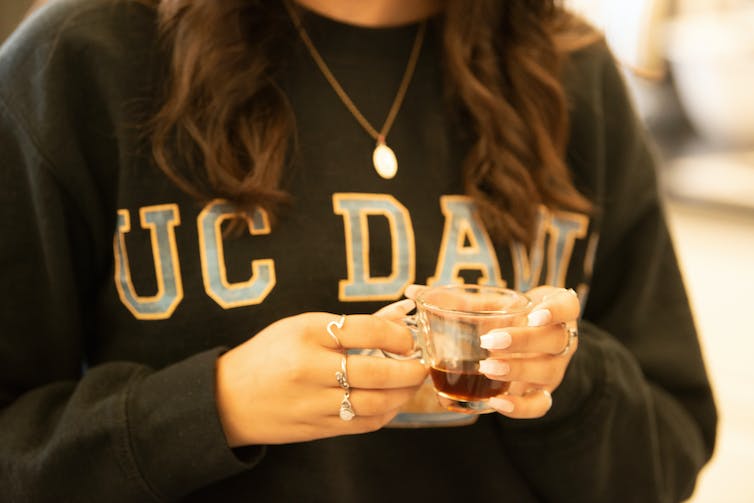 A student wearing a black UC Davis sweatshirt holds a glass cup of coffee
